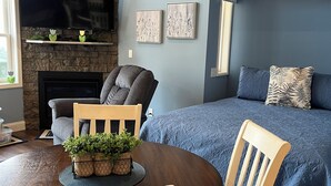 Ein gemütliches Wohnzimmer mit Kamin, einer bequemen Couch, einem Esstisch mit Stühlen und einem großen Bett mit blauer Bettdecke.