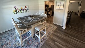 Uma sala de jantar com uma mesa de madeira e cadeiras brancas, cozinha ao fundo e um tapete estampado no chão.