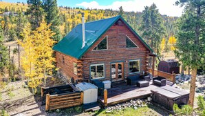 Una cabaña de troncos con techo verde, una terraza con mobiliario exterior y vistas a un área boscosa.