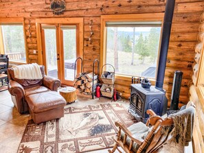 A cozy cabin interior with a leather chair, a wooden rocking chair, a fireplace, and a large window offering a view of the forest.