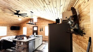 Un intérieur de chalet chaleureux avec un îlot central en pierre, un grand réfrigérateur, un ventilateur de plafond et des murs en bois.