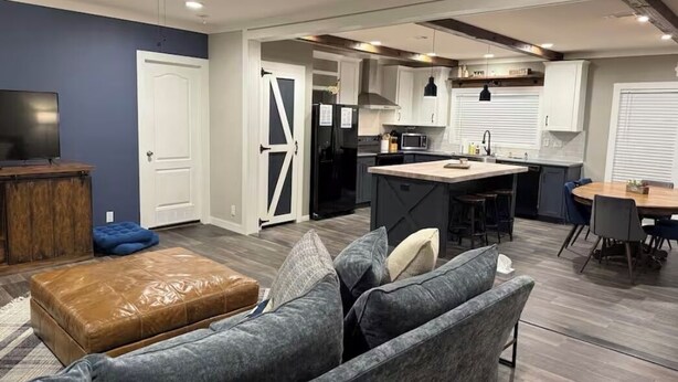 Una cocina moderna con una isla central, un comedor con una mesa redonda y una sala de estar con un sofá gris y un otoman marrón.