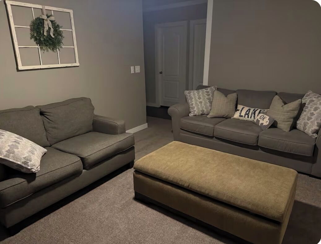 A living room with two grey sofas, a beige ottoman, and a wall-mounted wreath.