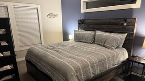 Un dormitorio con una cama de madera oscura, ropa de cama gris, una mesita de noche con lámpara y una ventana con persianas.