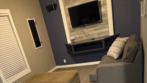 Una sala de estar con un sofá gris, un reposapiés marrón y un sillón gris. Un televisor de pantalla plana está montado en la pared sobre un panel de madera decorativo.