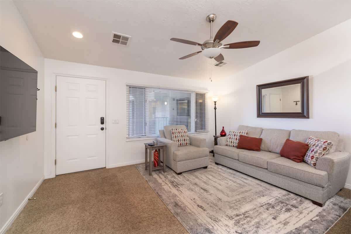 A living room with a ceiling fan, a large window with blinds, a sofa set, and a flat-screen TV mounted on the wall.
