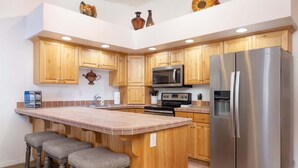 A kitchen with wooden cabinets, a stainless steel refrigerator, a microwave, and a bar area with stools. - Las Palmas Resort #1638 | All-Inclusive Amenities (Saint George)