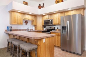 A kitchen with wooden cabinets, a stainless steel refrigerator, a microwave, and a bar area with stools. - Las Palmas Resort #1638 | All-Inclusive Amenities (Saint George)