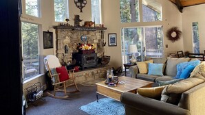 A cozy living room with a stone fireplace, large windows, and a high wooden ceiling.