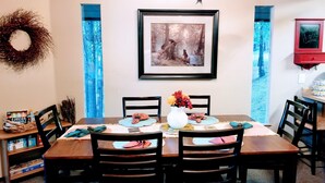 A dining room with a wooden table set for four, surrounded by chairs. A framed picture hangs above the table, and a red cabinet with a 'Welcome Home!' sign is visible to the right.