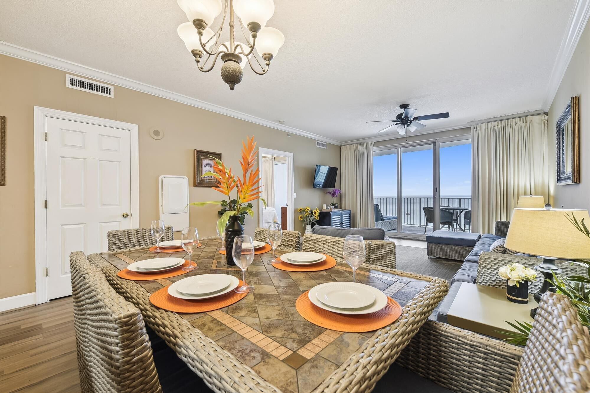 Un coin repas avec une grande table dressée pour quatre personnes, entourée de chaises en osier. Un lustre est suspendu au-dessus de la table, et un téléviseur est fixé au mur. La pièce donne sur un balcon d’où l’on peut admirer le ciel.