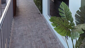 Un couloir extérieur abrité, doté d’un sol carrelé, de plantes en pot et offrant une vue sur les voitures garées au loin.