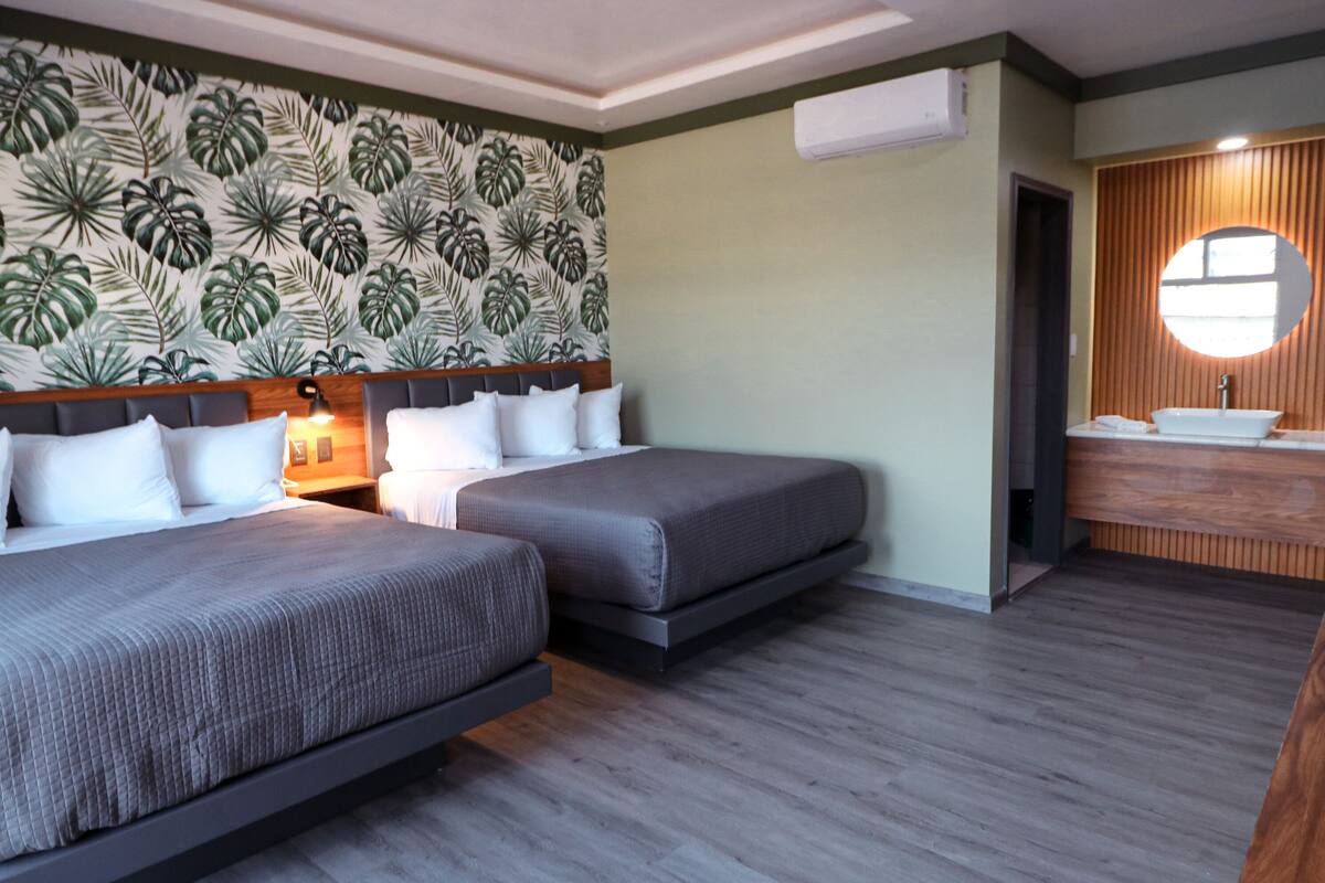 Habitación de hotel con dos camas, cabecero de madera, pared con estampado de hojas tropicales, espejo redondo sobre el lavabo y suelo de madera.