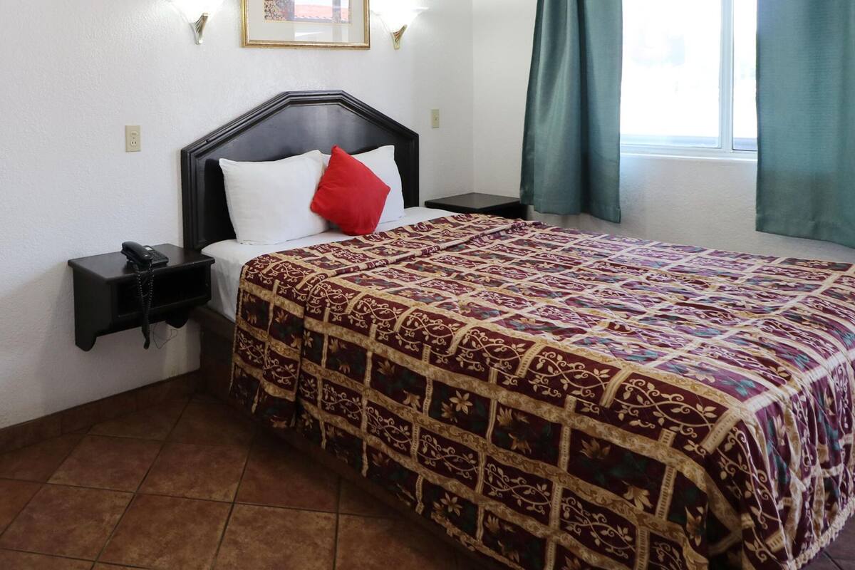 Habitación de hotel con cama, mesita de noche con teléfono, pequeño escritorio con lápiz y ventana con cortinas.