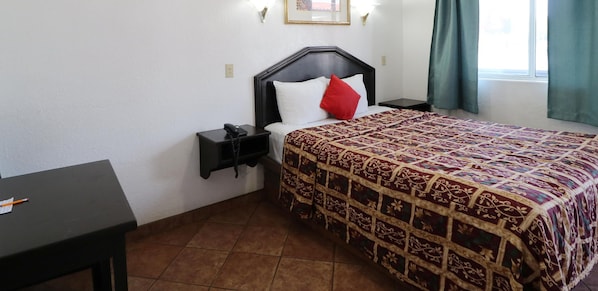 Habitación de hotel con cama, mesita de noche con teléfono, pequeño escritorio con lápiz y ventana con cortinas.