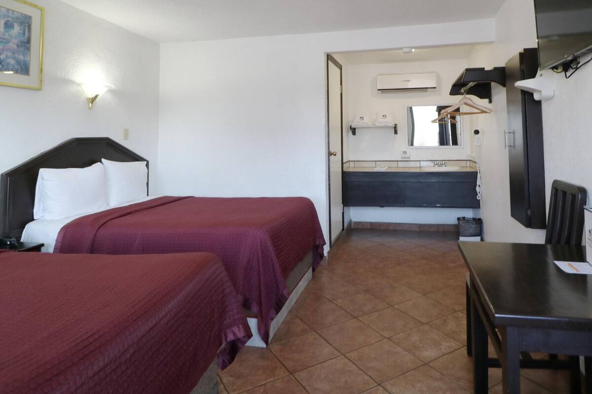 Habitación de hotel con dos camas, un escritorio, un televisor, un baño con espejo y lavamanos, y un cuadro en la pared.