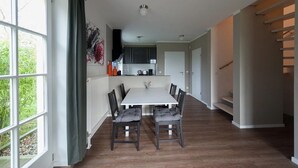 A spacious dining area with a white table and dark chairs, a kitchen in the background, and a view of greenery through large windows. - Naturferienhaus 2 - Naturferienhaus auf der Insel Rügen (Poseritz)