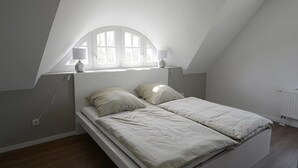 A bedroom with a sloped ceiling, a bed with two pillows, a window with a lamp, and a radiator. - Naturferienhaus 2 - Naturferienhaus auf der Insel Rügen (Poseritz)