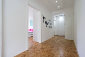 Um corredor com piso em padrão herringbone, paredes brancas e uma porta que dá para um quarto com cama e almofadas cor-de-rosa.