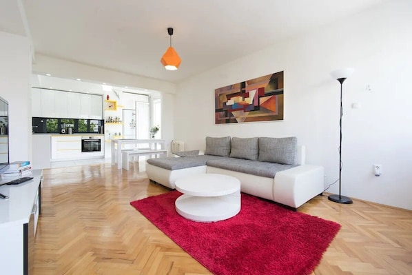 Uma sala de estar moderna com um sofá branco, um tapete vermelho e uma cozinha ao fundo.