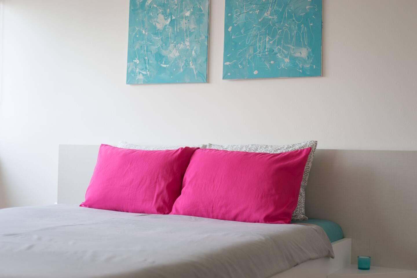 A bed with white bedding and two bright pink pillows, a teal wall art piece above, and a teal candle on the bedside table.