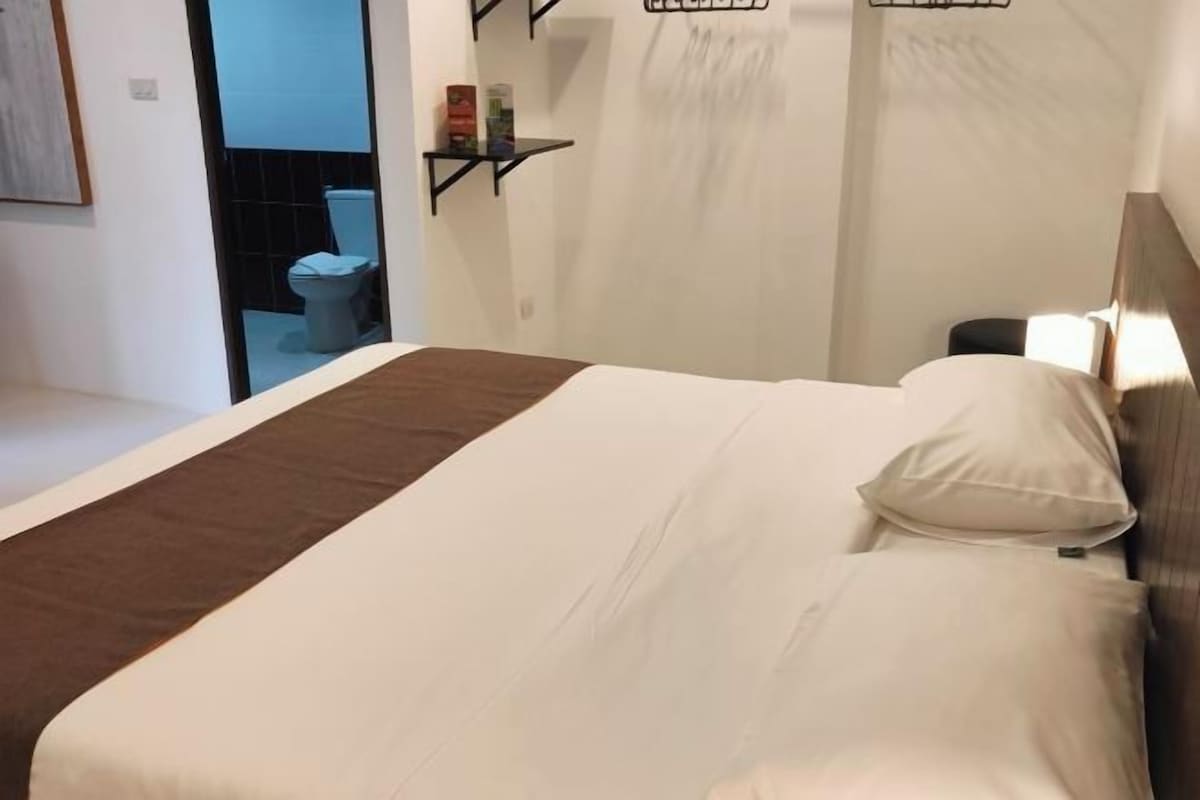 A hotel room with a bed, a bedside table, and a small chair. There is a window with a view of the outside.