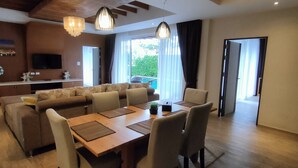 A modern living room with a sofa set, dining table, and chairs. There is a kitchen area with a refrigerator and a flat-screen TV. The room has large windows with curtains and a view of greenery outside. - Phuket Jungle Experience Resort (Karon)