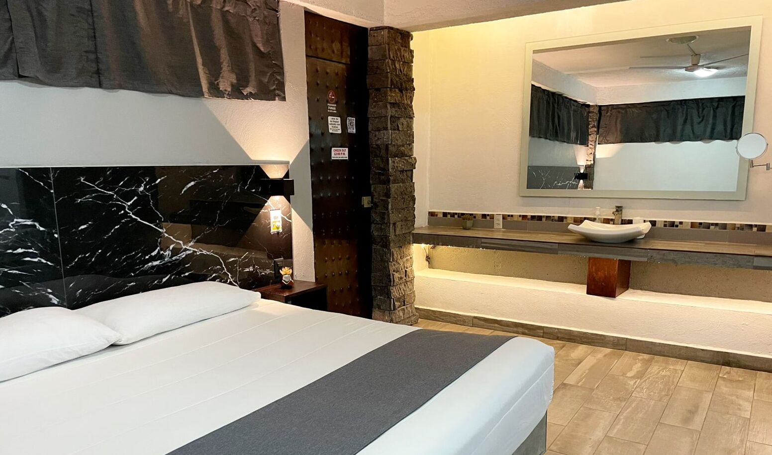 A hotel room with a bed, a nightstand, a bathroom with a sink and mirror, and a stone wall.