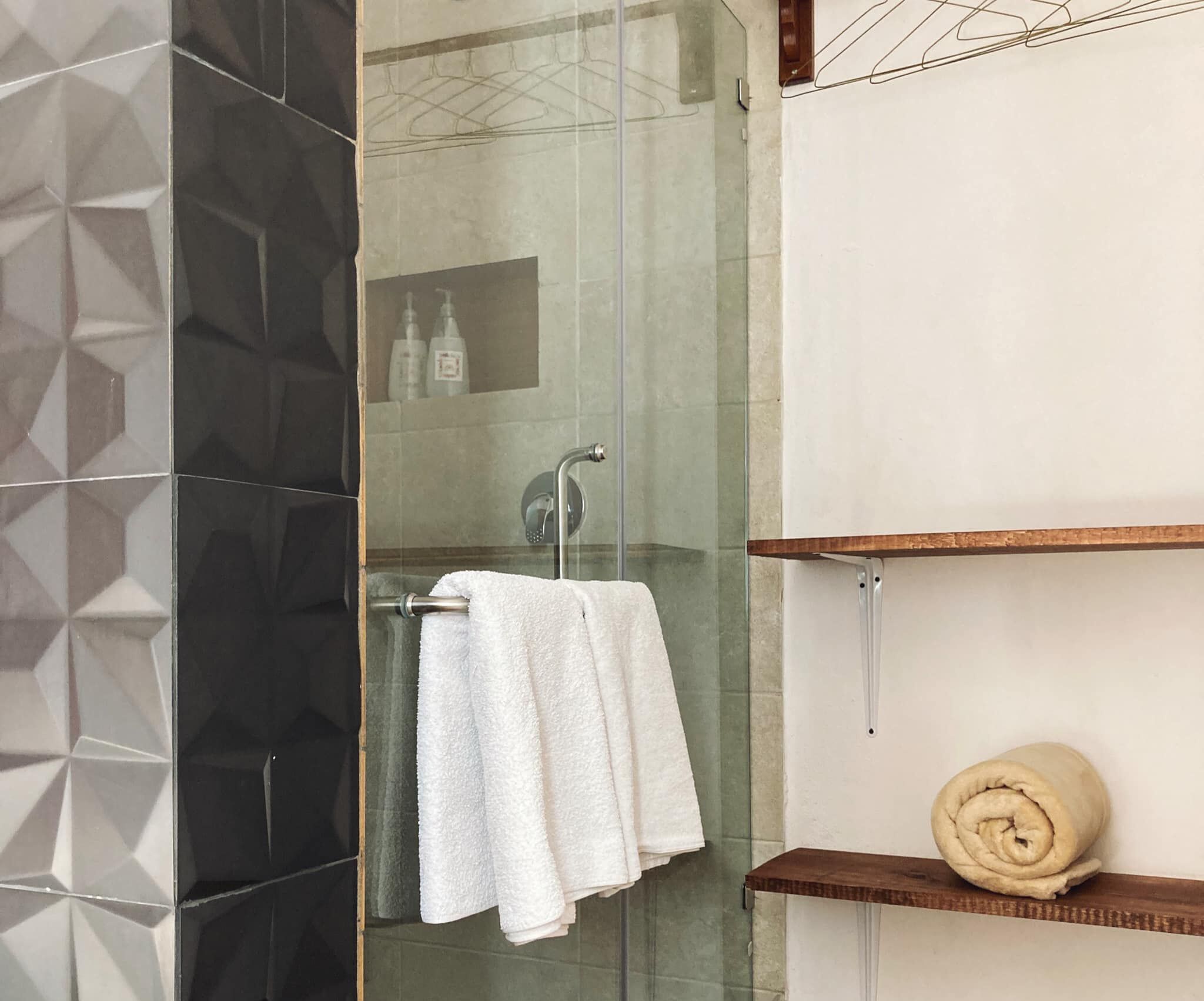Un baño moderno con cabina de ducha de vidrio, una toalla blanca colgada en un perchero y un estante de madera con una toalla enrollada.