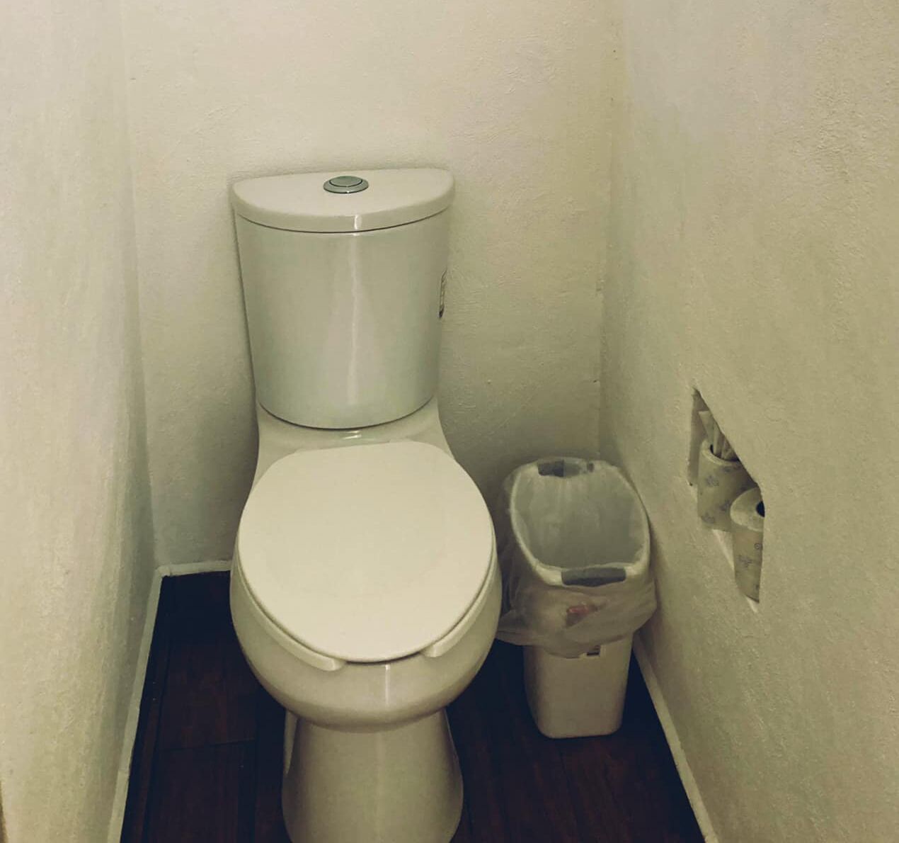 Un baño pequeño y blanco con un bote de basura a su lado en una habitación estrecha de paredes lisas.