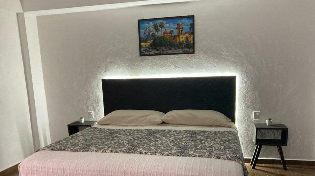 A bedroom with a bed, two bedside tables, a nightstand, a painting on the wall, and a headboard with built-in lighting.