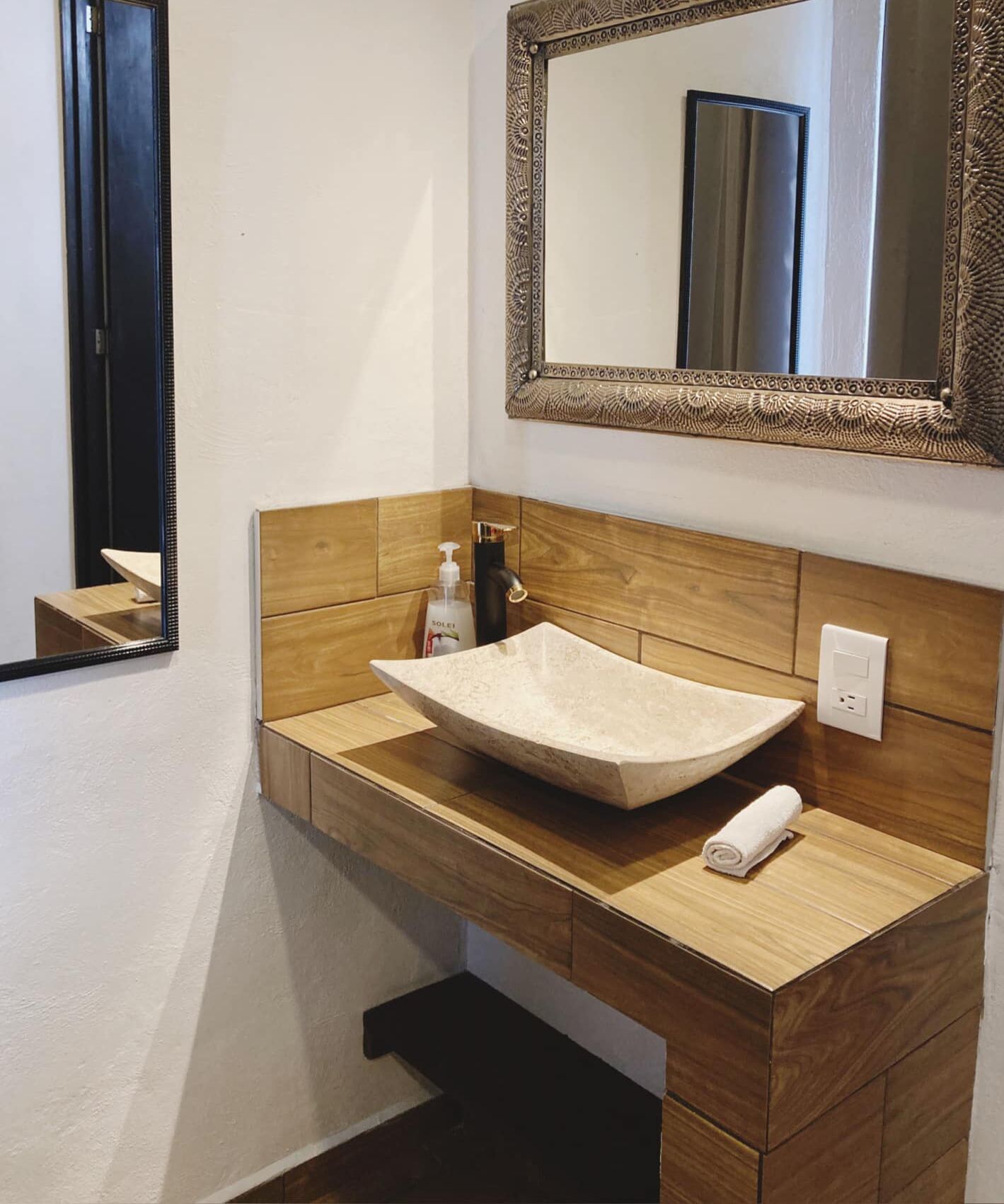 A bathroom with a wooden vanity, a beige sink, a mirror with an ornate frame, and a bottle of hand soap.