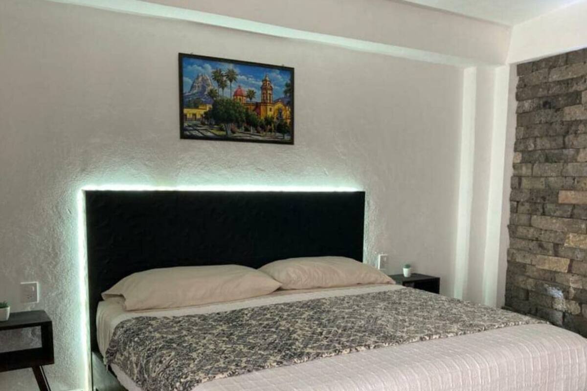 A bedroom with a bed, a headboard, a nightstand, a wall-mounted painting, and a brick wall.