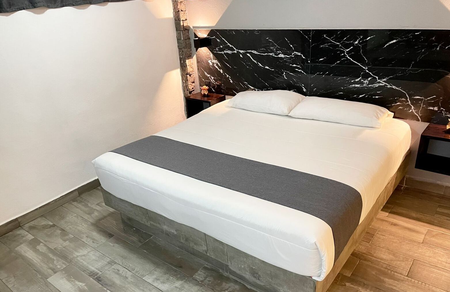 Un lit bien fait, recouvert d’une couvre-lit blanche et grise, avec une tête de lit à motifs de marbre et une table de chevet en bois surmontée d’une lampe.
