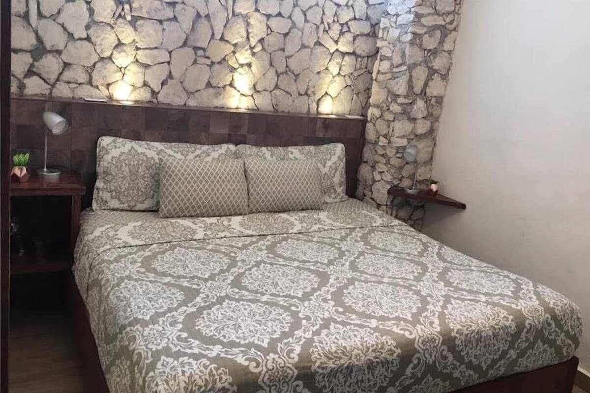A bedroom with a stone-patterned headboard, a patterned bedspread, and a bedside table with a lamp and a small plant.