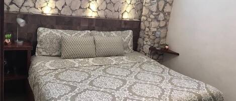 A bedroom with a stone-patterned headboard, a patterned bedspread, and a bedside table with a lamp and a small plant.