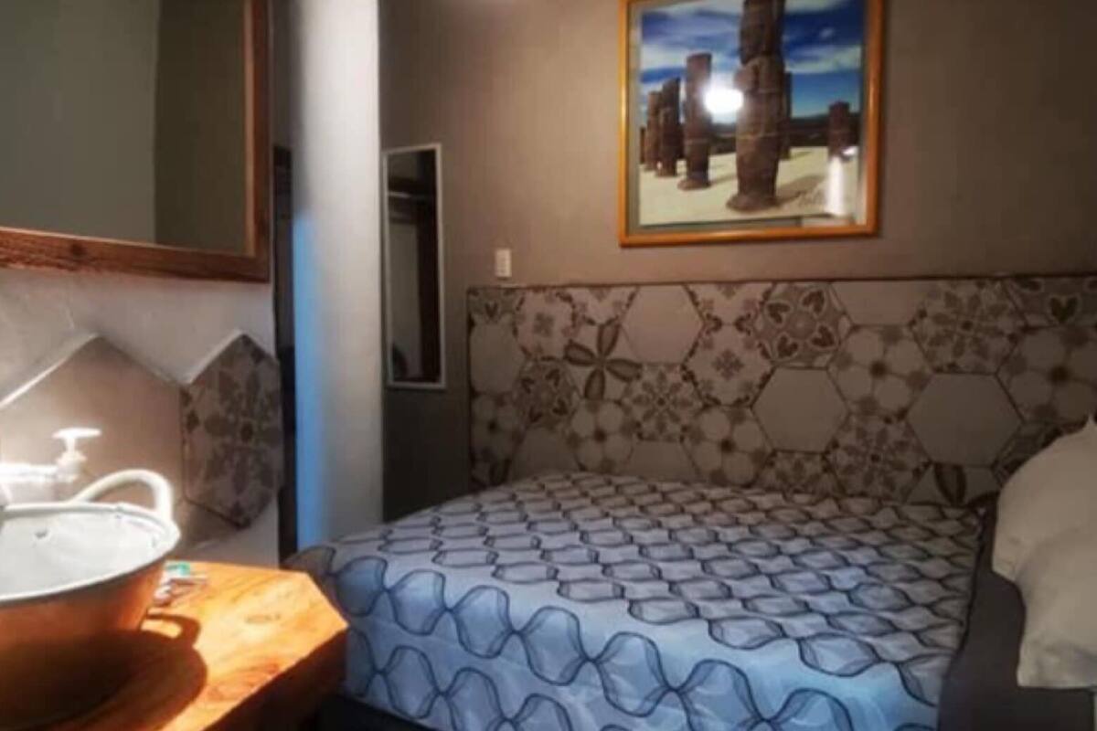 A bedroom with a patterned bedspread, a ceiling fan, a framed painting, and a wooden nightstand with a lamp.