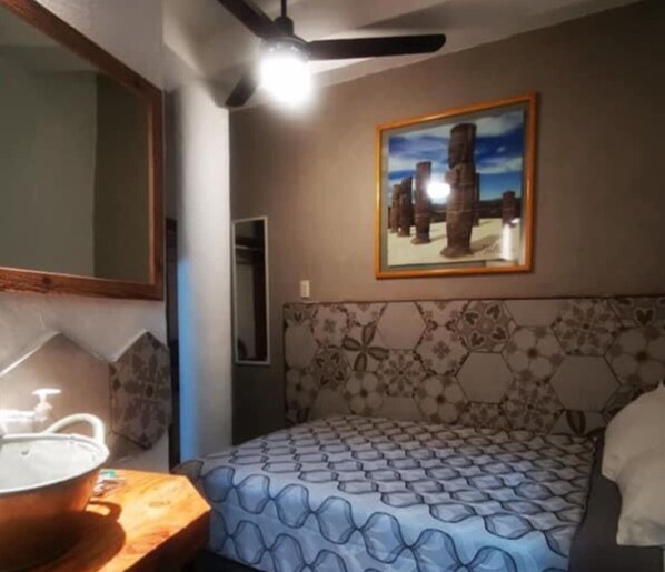Un dormitorio con una colcha estampada, un ventilador de techo, un cuadro enmarcado y una mesita de noche de madera con una lámpara.