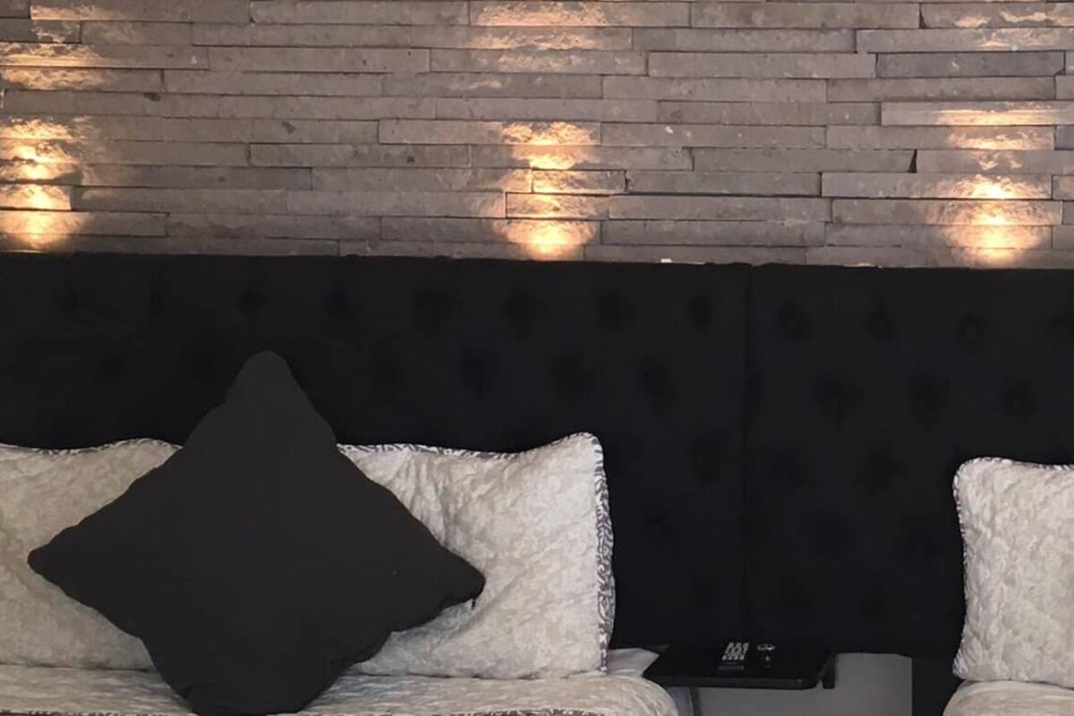A hotel room with a brick wall, two beds with white and dark pillows, and a small table with a clock.