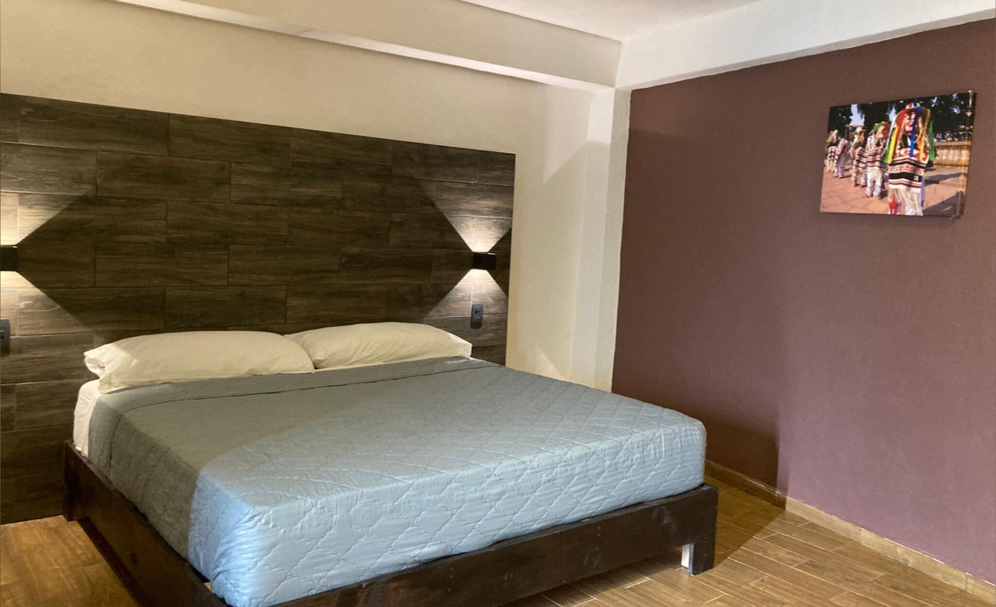 A bedroom with a wooden headboard, a bed with a grey quilt, and a wall-mounted picture.