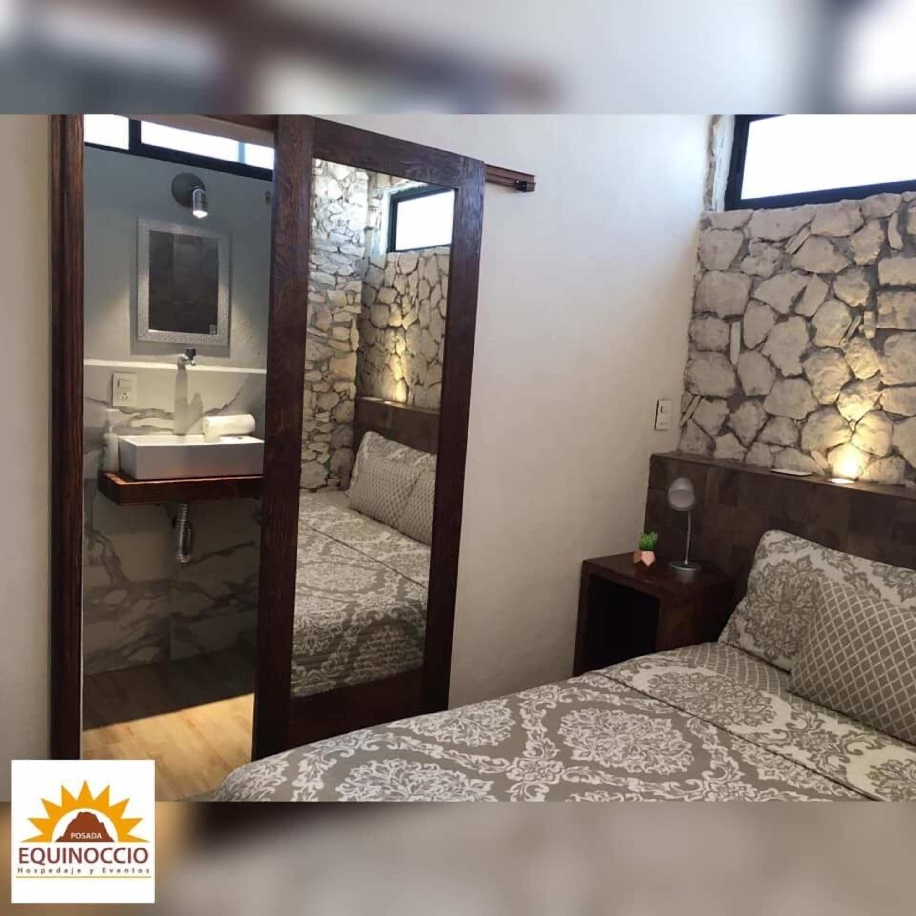 A bedroom with a stone wall, a wooden bed frame, a mirror, a bedside table, and a nightstand.