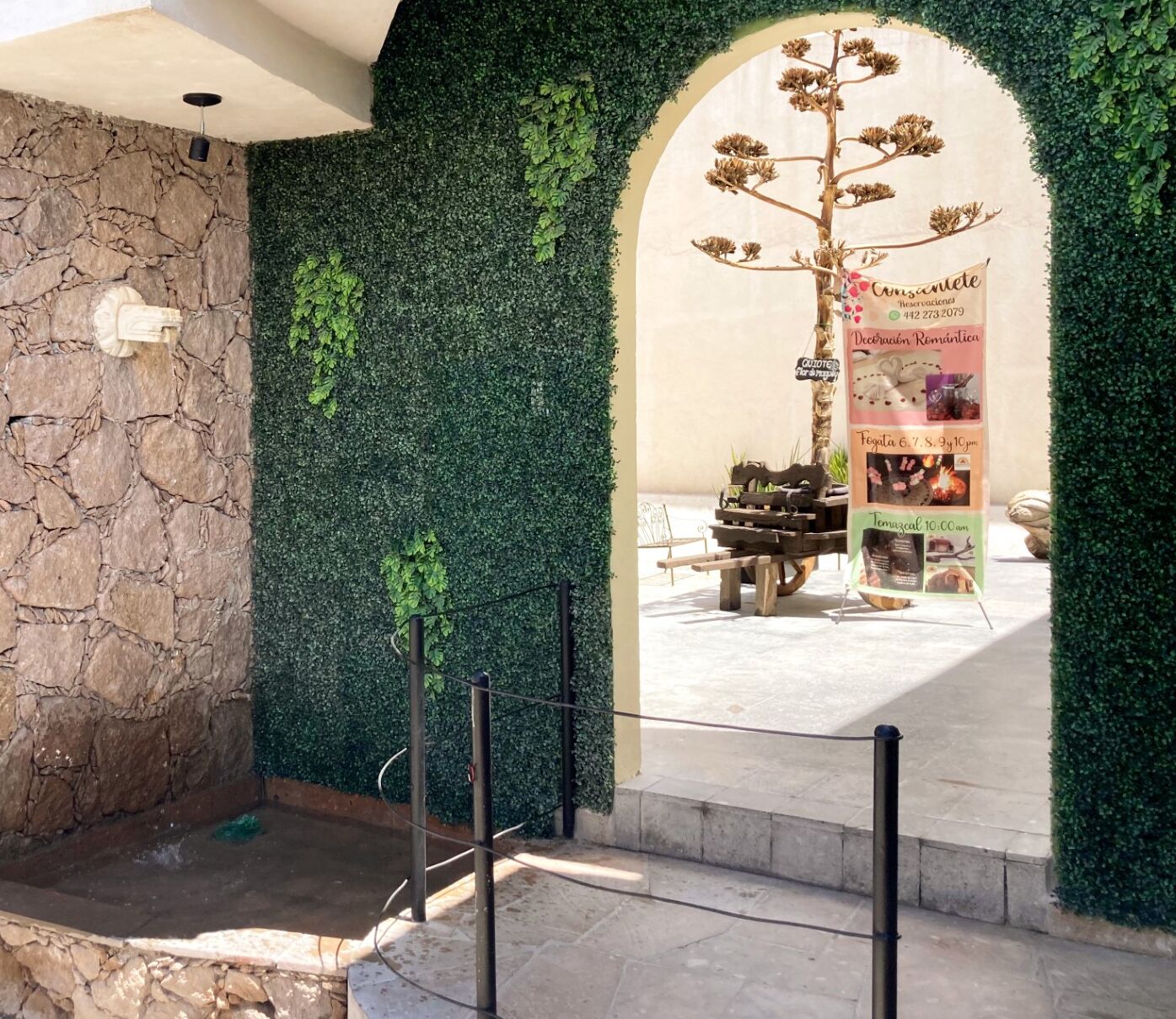 A stone wall with a green hedge, an archway leading to a shop, and a wooden cart with items.