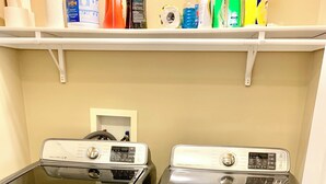 A laundry area with two stainless steel front-loading washers and dryers, a shelf above with cleaning supplies, and a white wall.