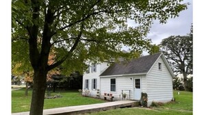 A small, single-story white house with a gabled roof, a front porch, and a small front yard with a tree and a pathway leading to the door. - Oak Meadow Farmhouse (Spring Valley)