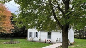 A white single-story house with a front porch, a small garden, and a tree with green leaves. - Oak Meadow Farmhouse (Spring Valley)