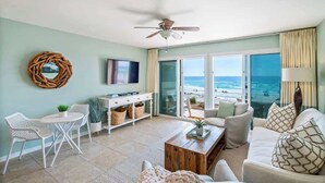 Un salon spacieux avec une grande fenêtre donnant sur la plage, un ventilateur de plafond et un canapé blanc.