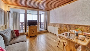 Ett mysigt vardagsrum med ett träpanelat tak, en soffa med ett mönstrat kuddöverdrag, en tv-apparat på ett träskåp och en matplats med ett bord dukat för två personer.