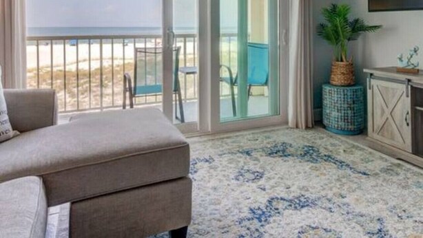 A room with a sofa, a potted plant, and a view of the beach through a sliding glass door.