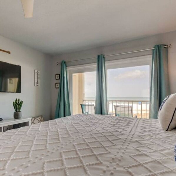 A bedroom with a large bed, a desk, a chair, a potted plant, and a window with curtains.