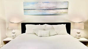 Ein ordentlich bezogenes Bett mit weißen Leinen und Kissen, zwei Nachttischlampen und ein Strandbild über dem Bett.